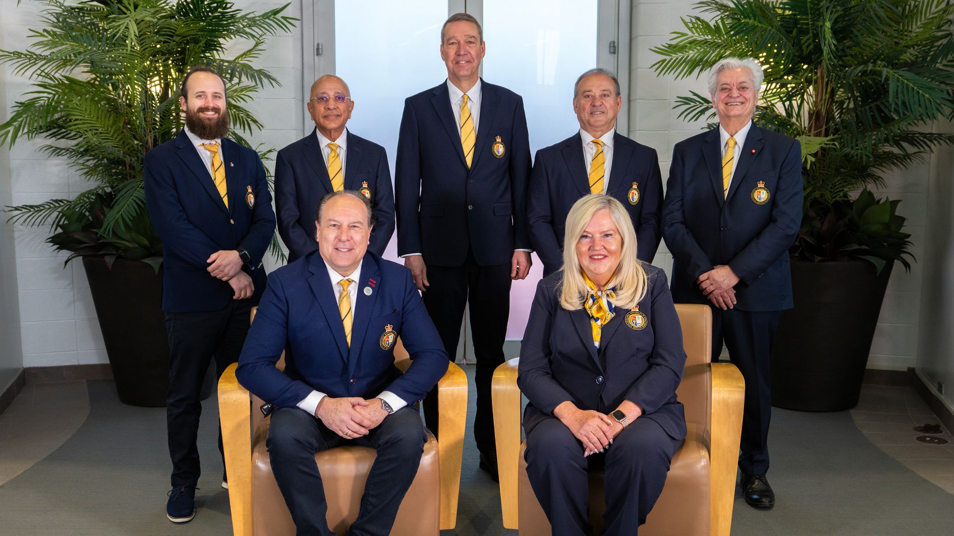 Seven people in matching navy blazers with embroidered crests and yellow striped ties pose for a formal group photo. Five stand in the back row between two large potted palms, and two sit in tan armchairs in front.